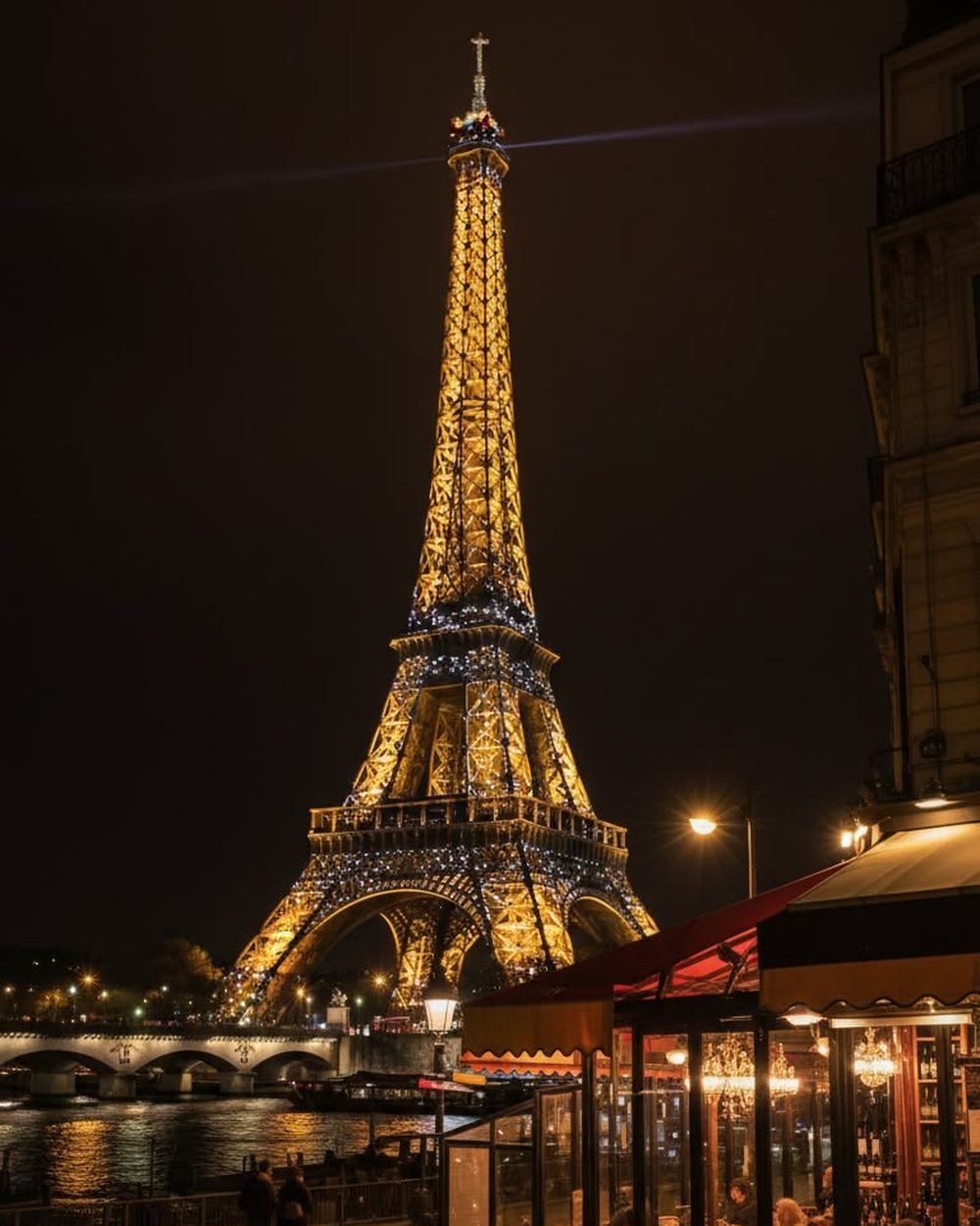 The Eiffel Tower lit at night, seen from a riverside cafe terrace.